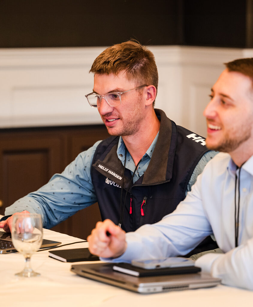 Two men sit at a conference table in a meeting, one wearing a dark vest and glasses, working on a laptop with notebooks nearby.