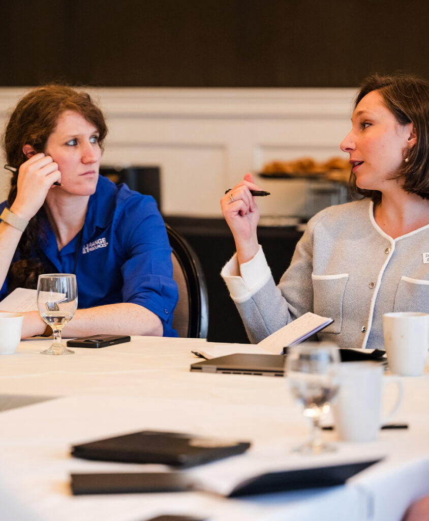 Two women converse at a table during a meeting, one in a blue shirt listening with a pen near her ear while the other speaks and gestues with a pen.